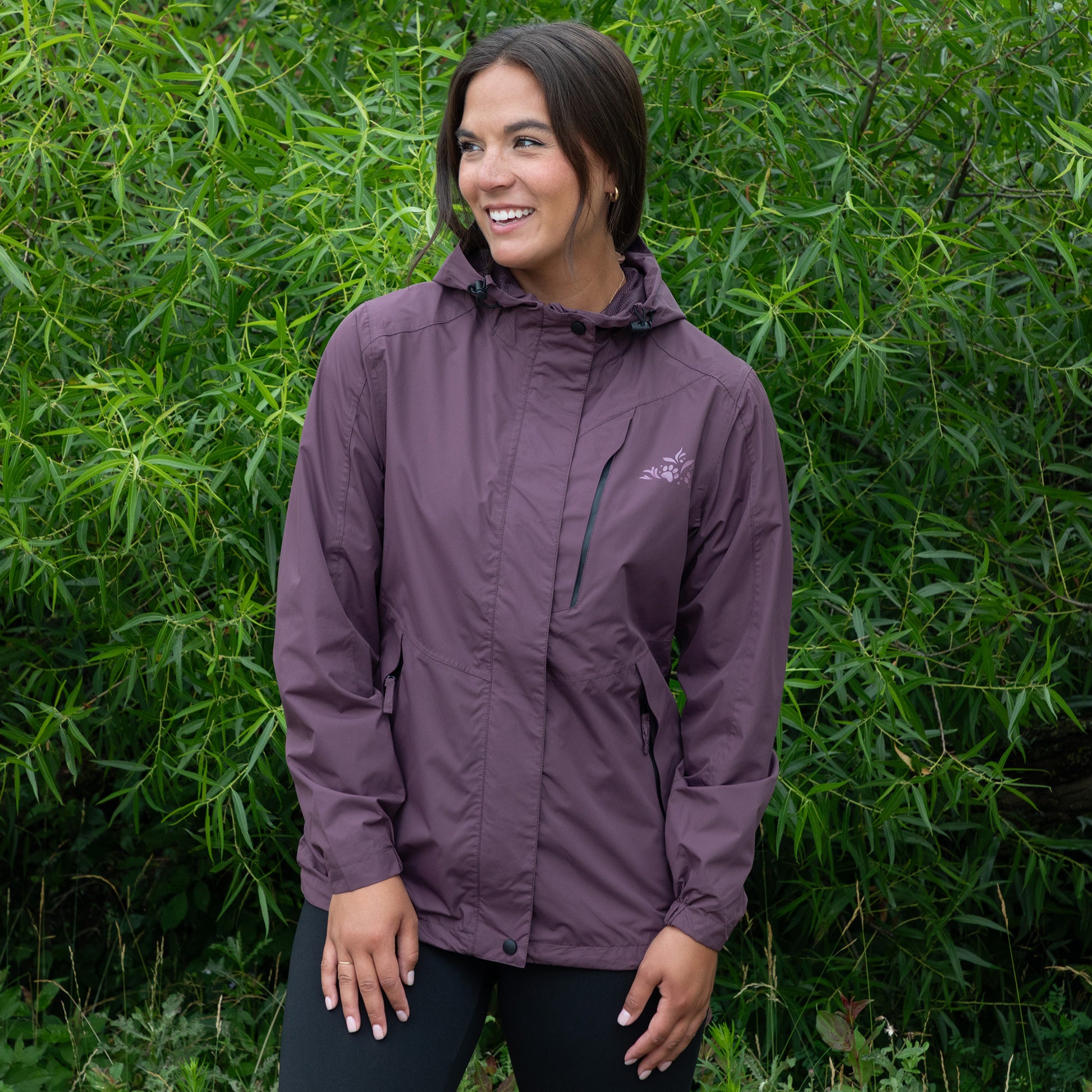 Woman wearing a purple paw rain jacket standing in front of green foliage outside.