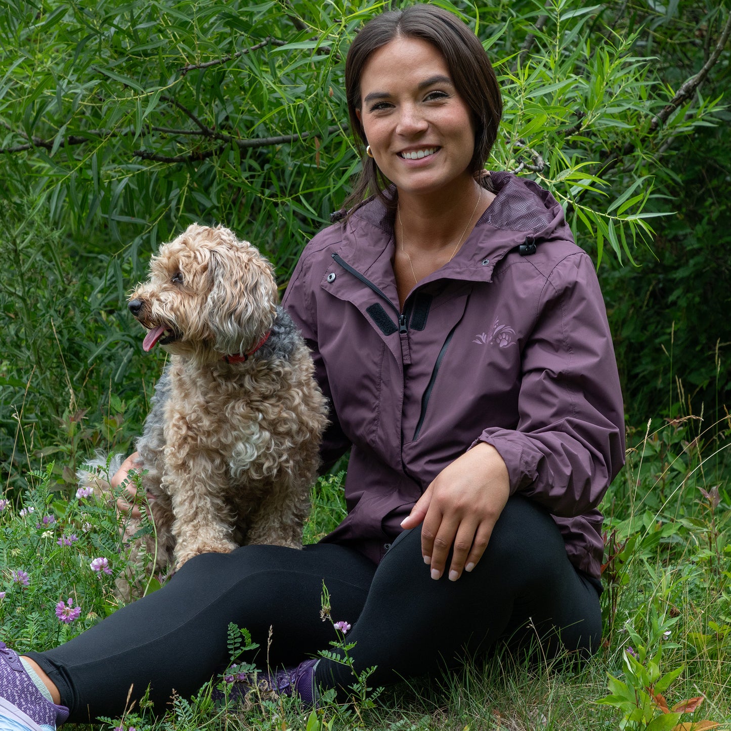 Woman in a purple rain jacket sitting with a dog in a grassy area with greenery.