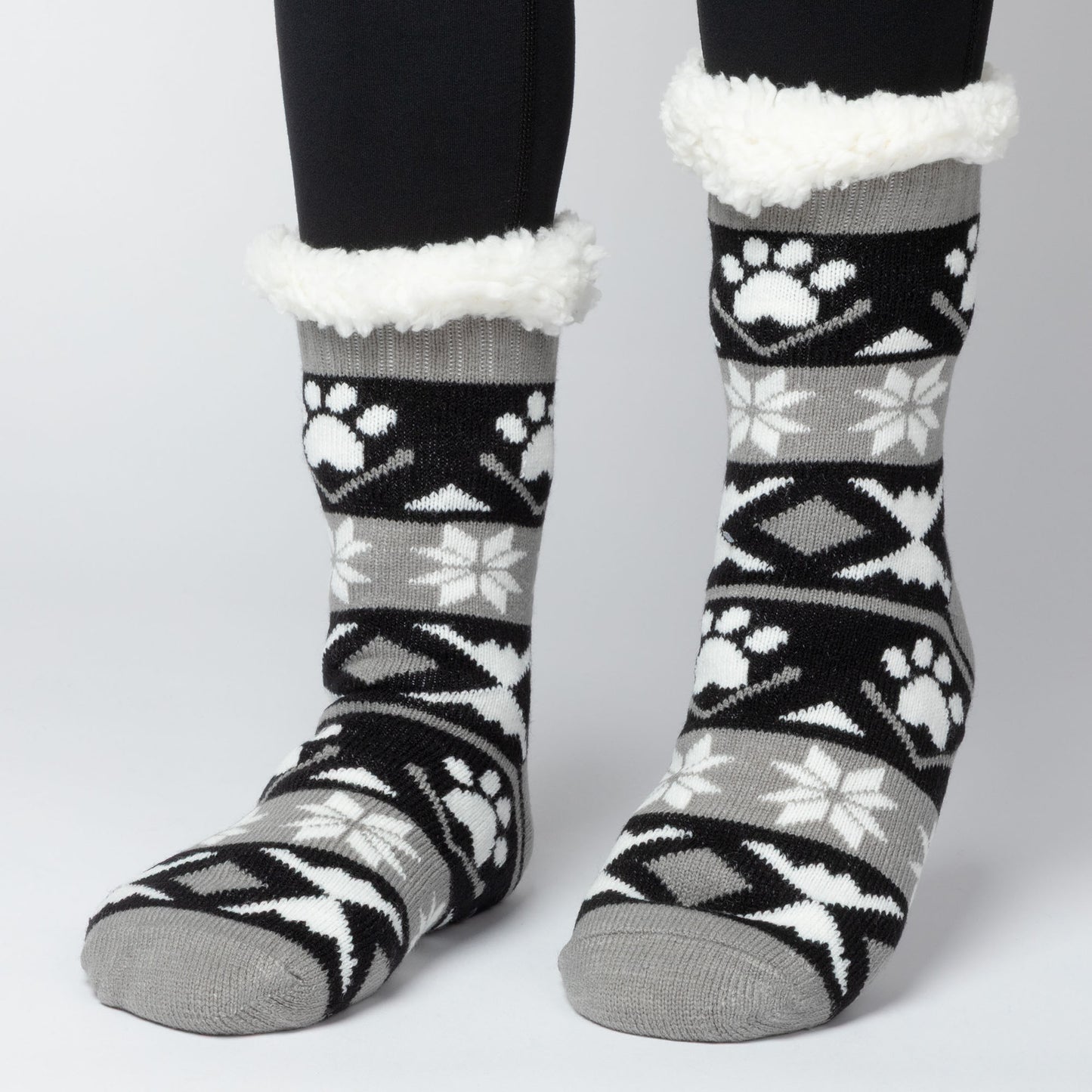 Model wearing Pair of gray, black white Sherpa Fleece Lined Paw Print Cabin Slipper Socks with sherpa trim on a light gray background alpine fair isle paws black