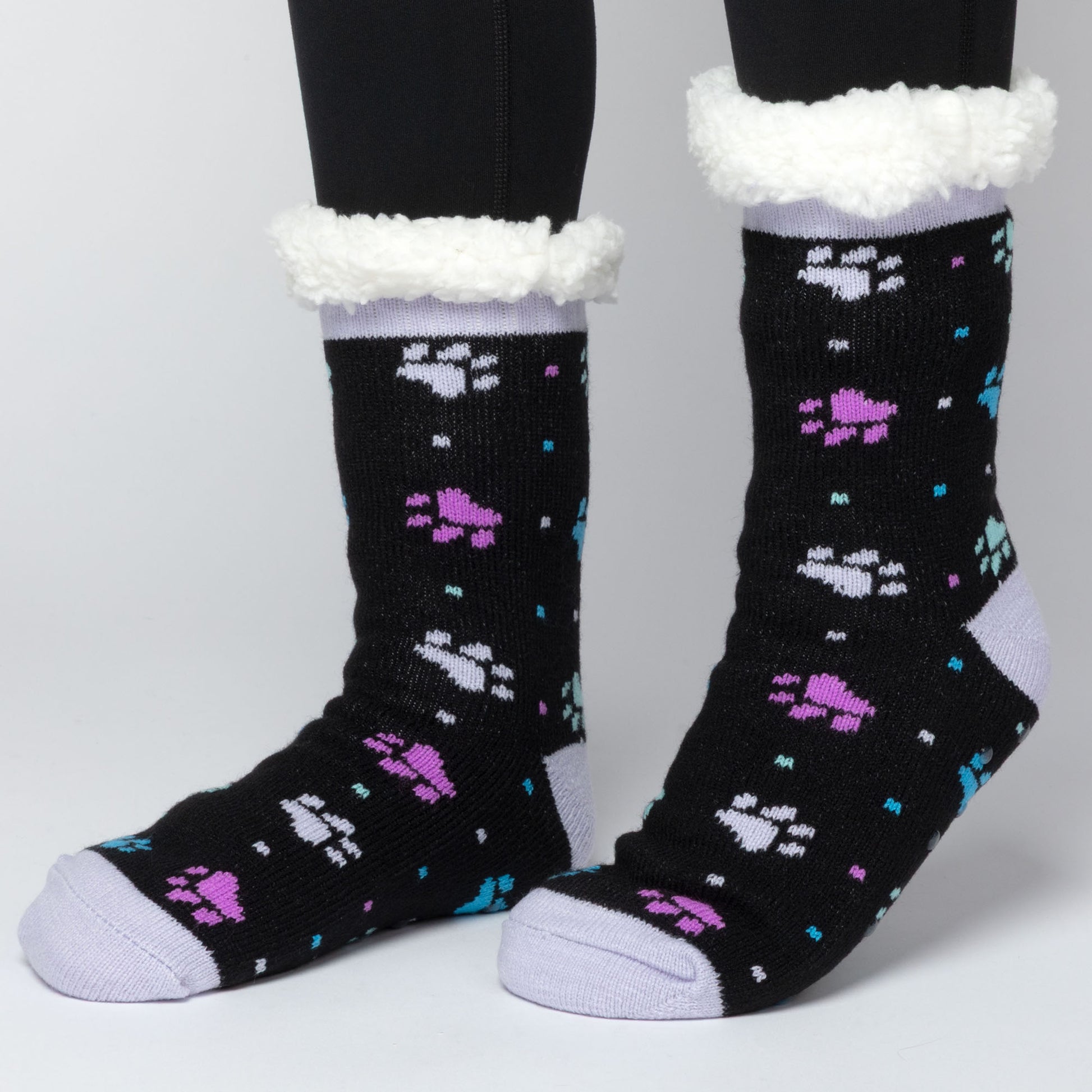 model wearing Pair of black with purple and blue polka dot paws Sherpa Fleece Lined Paw Print Cabin Slipper Socks with sherpa trim on a light gray background polka dot paws 