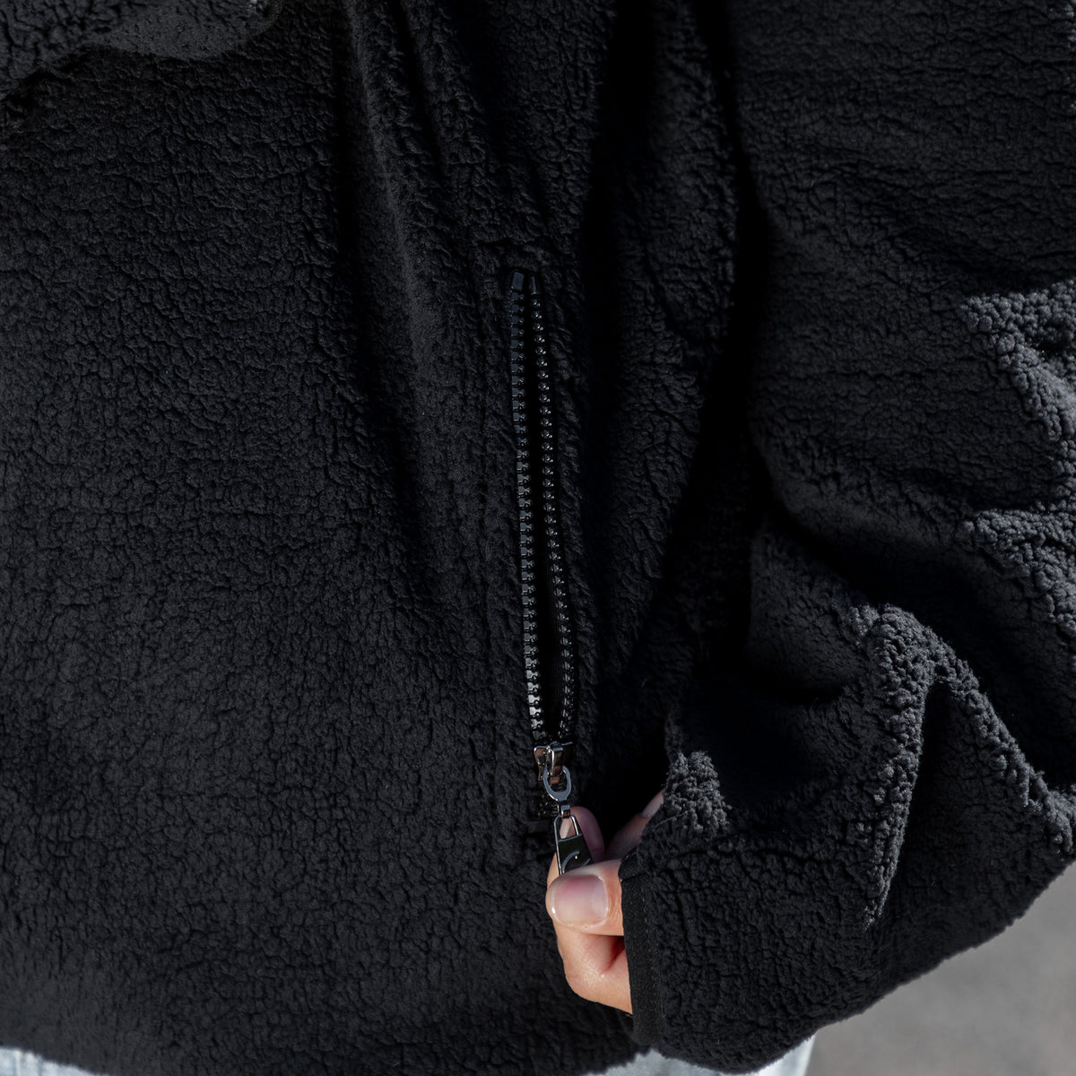Close-up of a black sherpa fleece jacket with a black zipper being pulled by a hand.