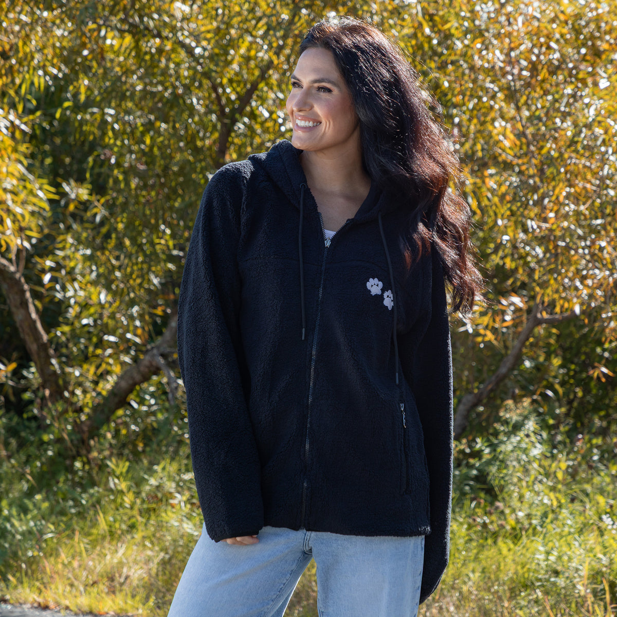 A woman wearing a black fleece zip-up hoodie with a small white paw print logo on the left chest, standing outdoors in front of a background of green and yellow autumn trees.
