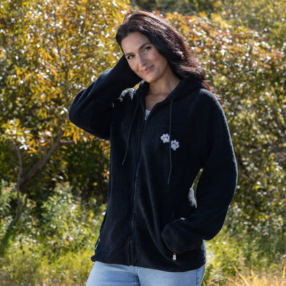 A woman wearing a black fleece zip-up hoodie with white embroidered paws on the chest, standing outdoors in front of a blurred green and yellow foliage background.