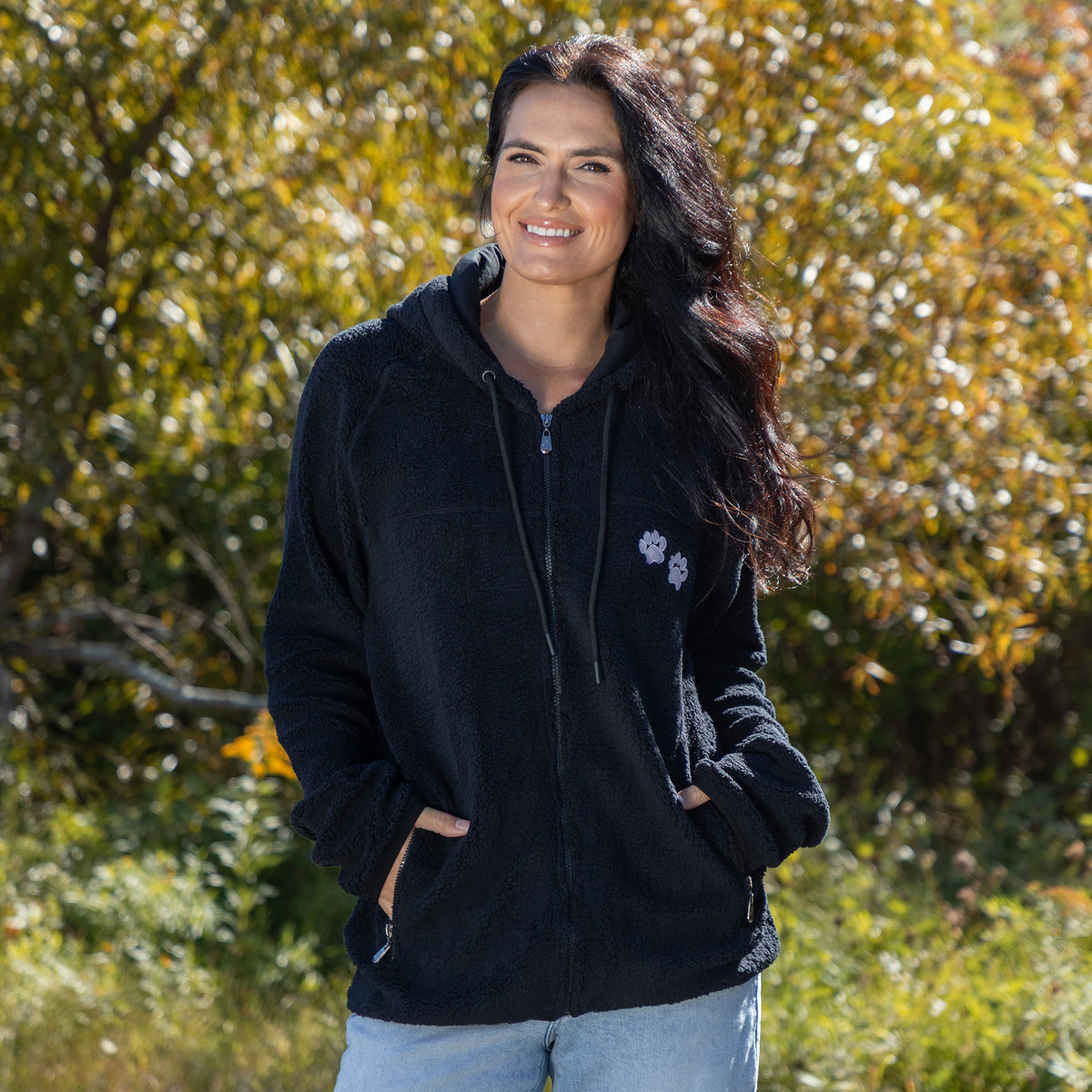 Smiling woman in a black fleece hoodie, surrounded by greenery and autumn foliage.