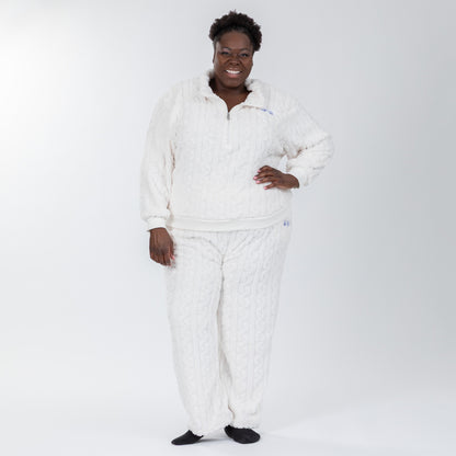 Woman wearing a white, fleece, cable-knit, quarter-zip, long-sleeve shirt with two light blue paw prints embroidered beneath the lapel. Her pants are part of the matching set and are also white with a cable-knit pattern and two light blue paw prints embroidered on the lefthand pocket.  Extended and plus size available. 