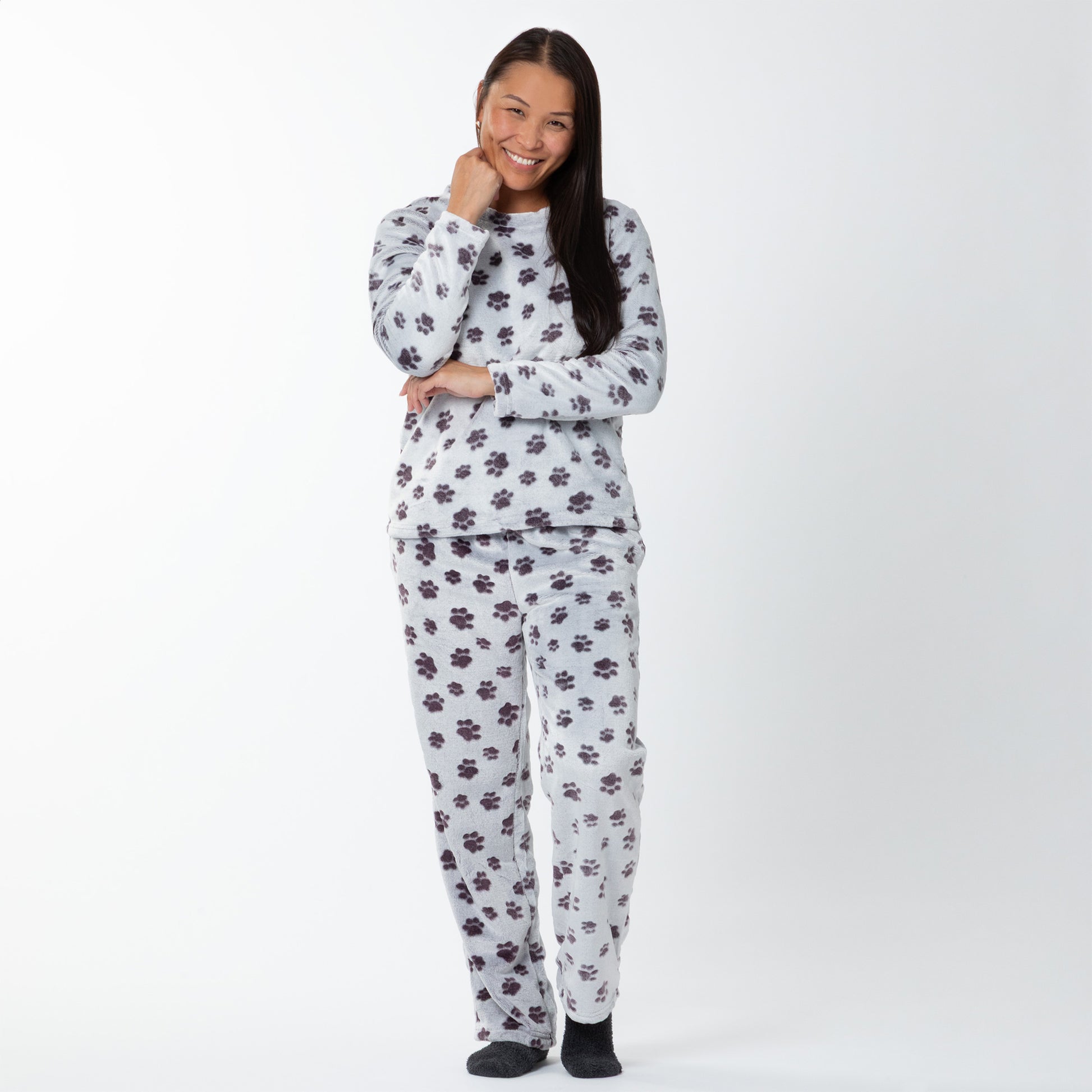 A woman wearing a matching set of fluffy, soft long-sleeve pajamas. The top and pants are light grey and patterned with purple paw prints. 