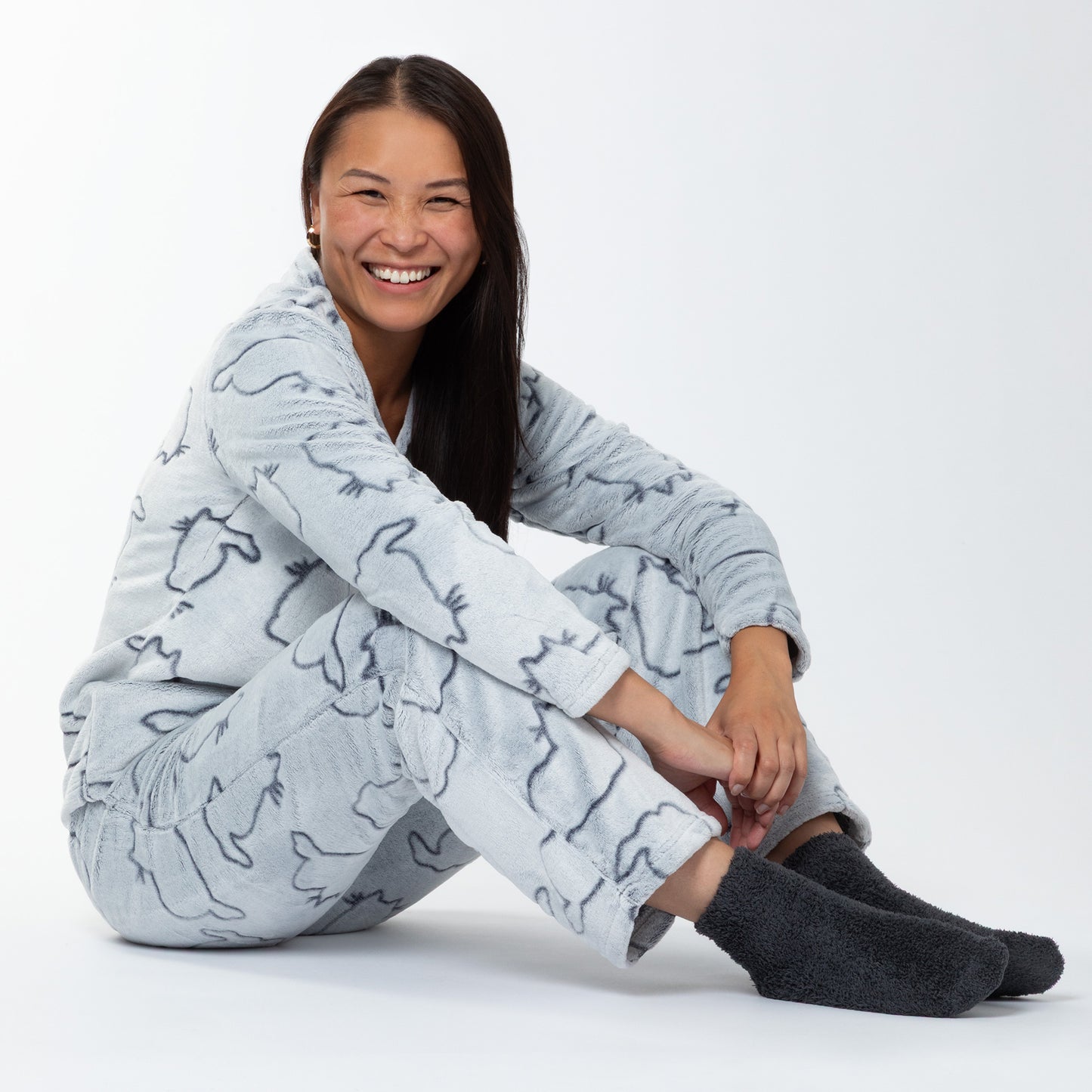 A woman sitting in a relaxed position. She is wearing a matching set of fluffy, soft long-sleeve shirt and pants pajamas. The top and pants are light grey and patterned with cute cat outlines. 