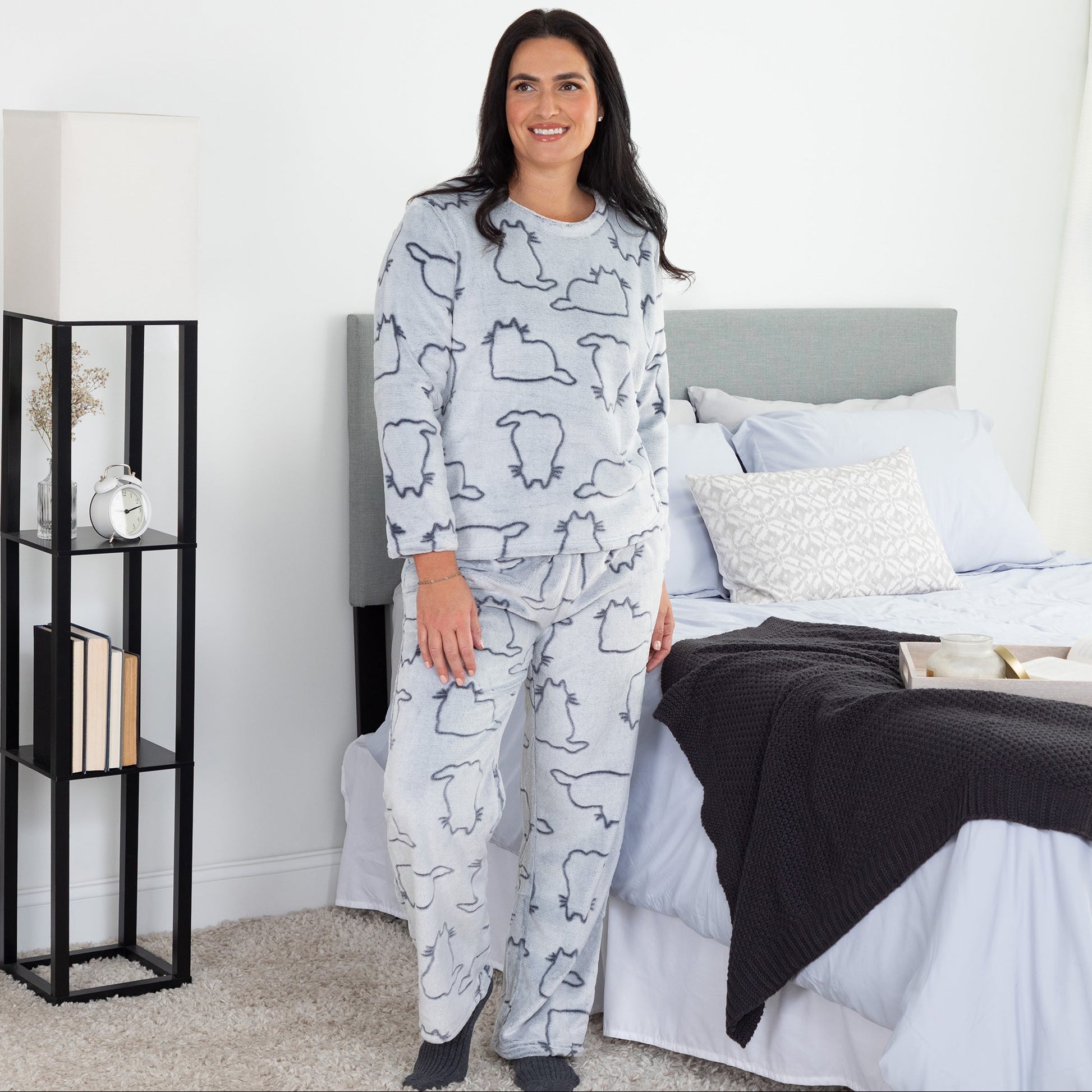 A woman standing in a cozy bedroom setting. She is wearing a matching set of fluffy, soft long-sleeve pajamas. The top and pants are light grey and patterned with cute cat outlines. 