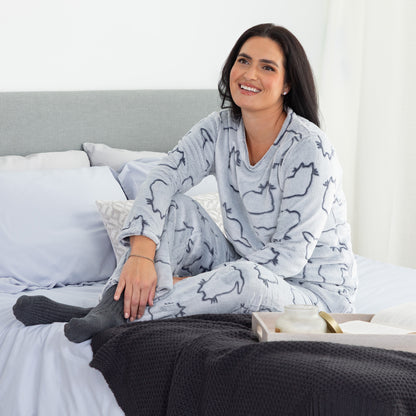 A woman lounging on a cozy bed. She is wearing a matching set of fluffy, soft long-sleeve pajamas. The top and pants are light grey and patterned with cute cat outlines. 