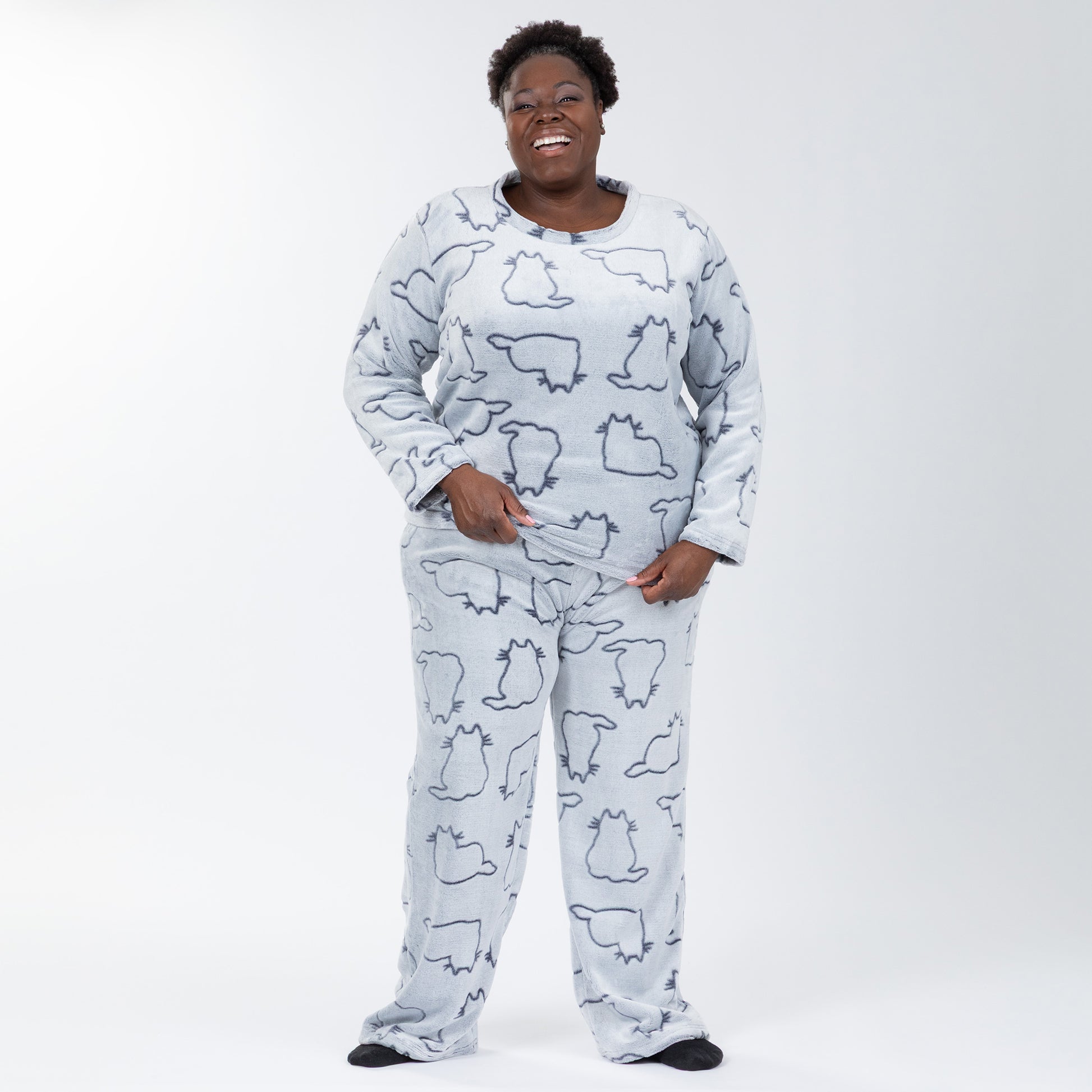 A woman wearing a matching set of fluffy, soft long-sleeve pajamas. The top and pants are light grey and patterned with cute cat outlines. Plus and extended size available.