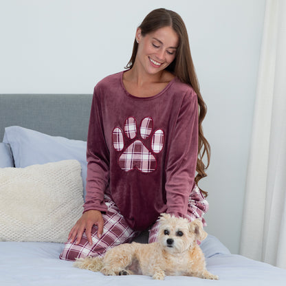 Woman sitting in cozy bed with her pet dog. She is wearing a velour pajama set— the top is a maroon long-sleeve shirt with a large plaid paw print. The pants are the same white and maroon plaid pattern as the paw print on the shirt. 