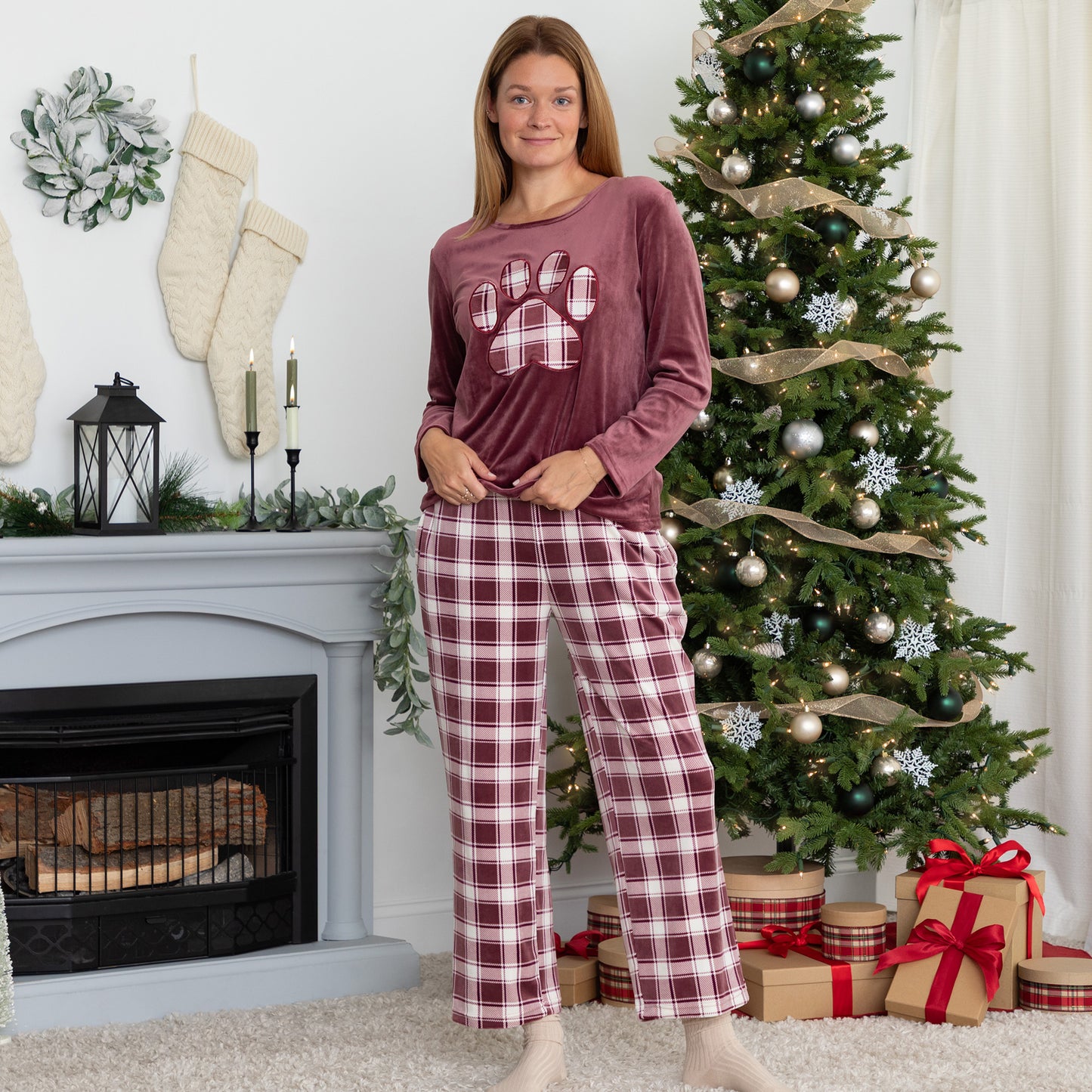 Woman standing in cozy holiday setting. She is wearing a velour pajama set— the top is a maroon long-sleeve shirt with a large plaid paw print. The pants are the same white and maroon plaid pattern as the paw print on the shirt. 
