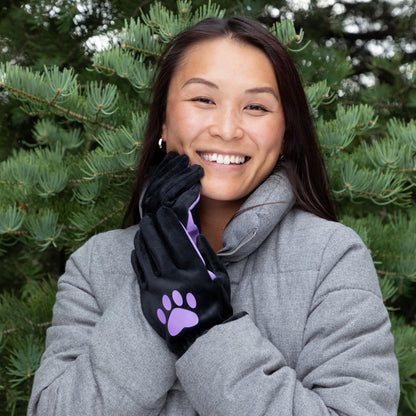 Woman wearing a gray coat and black gloves with a purple paw print, standing in front of green trees.