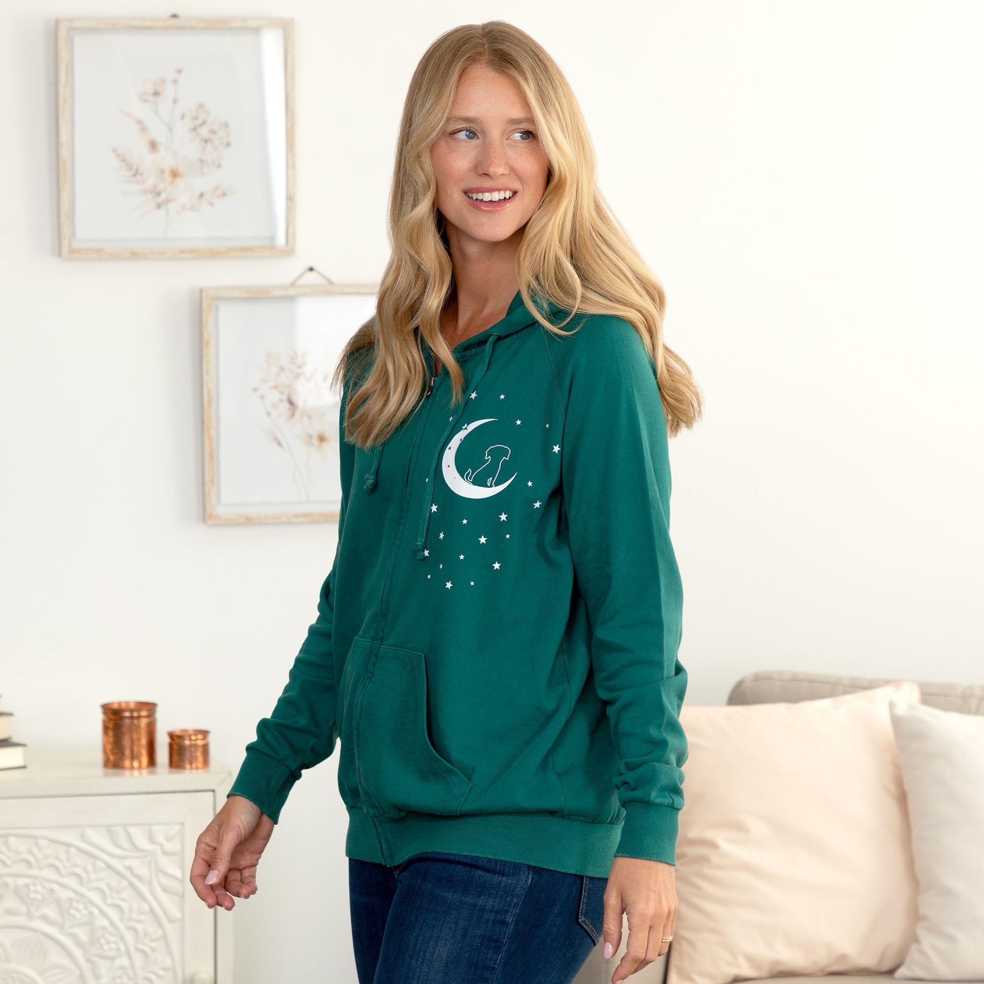 A woman wearing a dark teal green hoodie with a white crescent moon and stars graphic on the front, standing in a brightly lit living room.