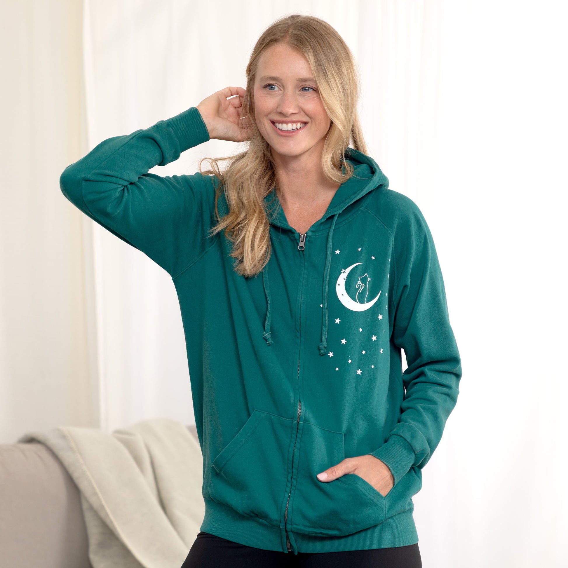 A woman wearing a teal green full-zip hoodie with a white crescent moon and stars graphic on the chest, posing in a brightly lit indoor setting.