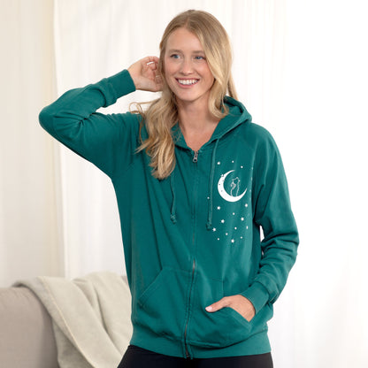 A woman wearing a teal green full-zip hoodie with a white crescent moon and stars graphic on the chest, posing in a brightly lit indoor setting.