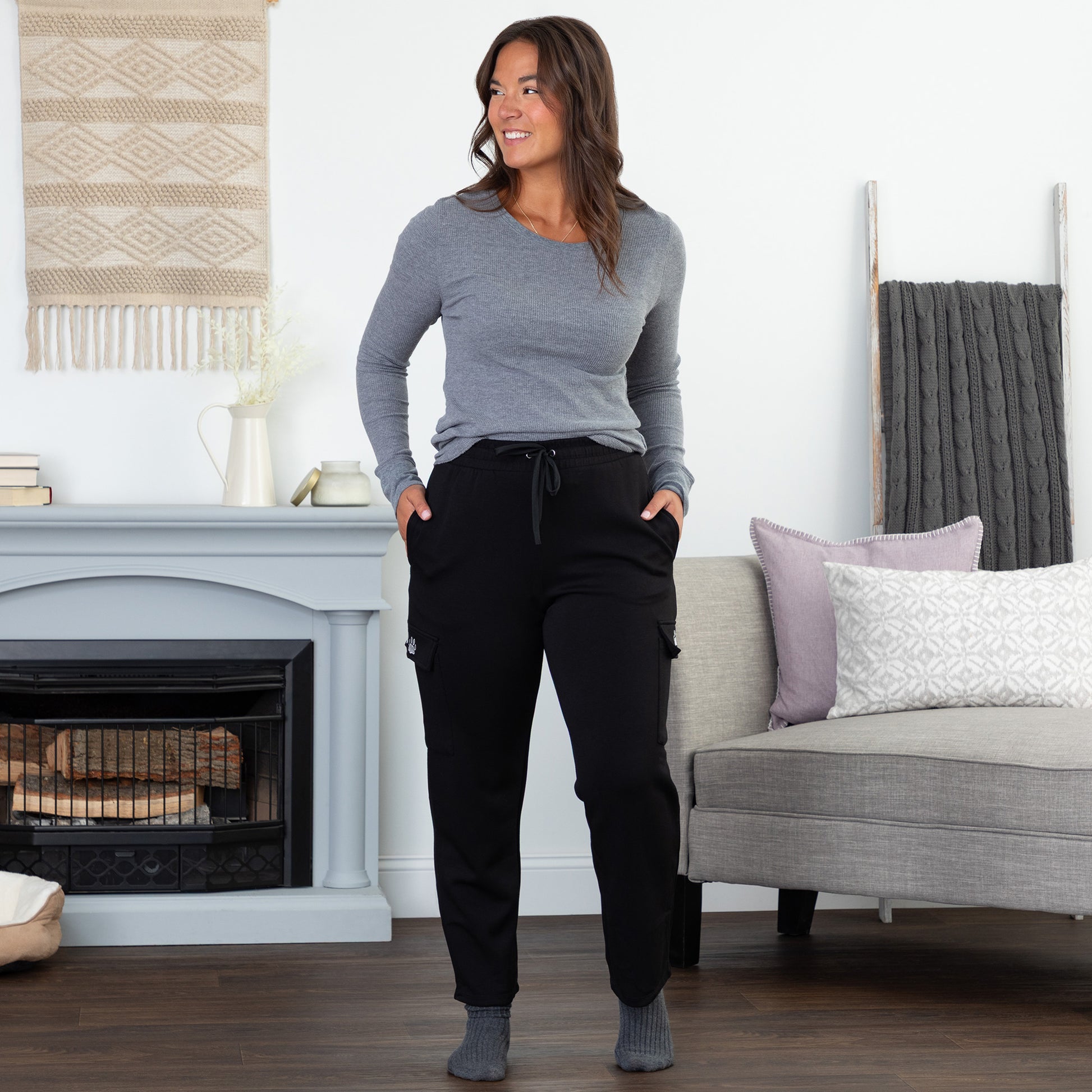 Woman standing in a living room wearing a gray long-sleeve top and black cargo pants.