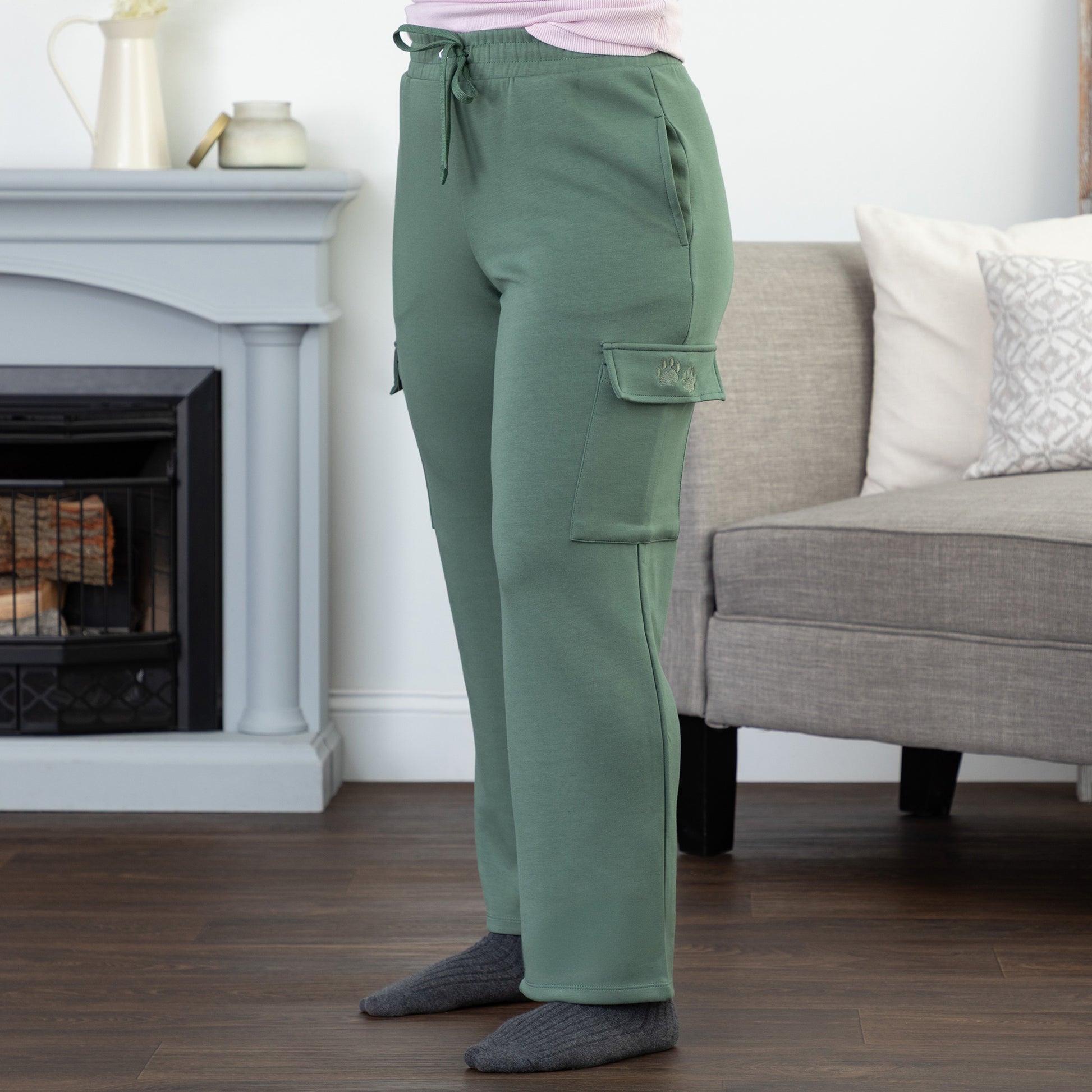 Person wearing green cargo pants with two embroidered paw prints on the pocket, standing in a living room setting.