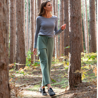 Woman walking through a forest wearing a gray long sleeve tee and green cargo pants with a drawstring waist and embroidered paws on the pocket.