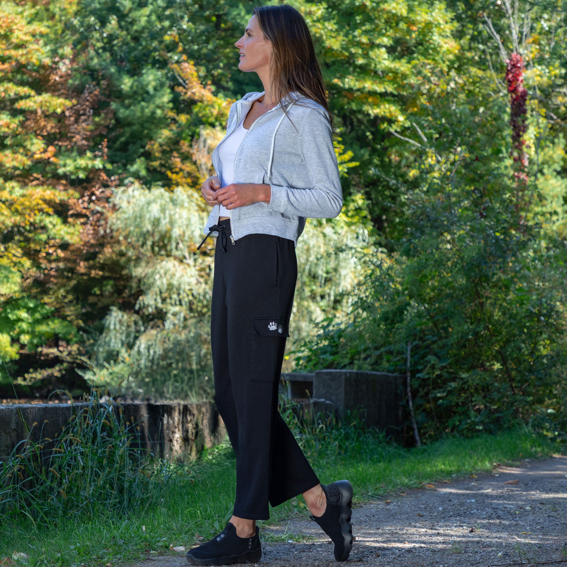 Woman walking outdoors in a natural setting with trees and greenery, wearing black cargo pants with two embroidered paw prints on the pocket.