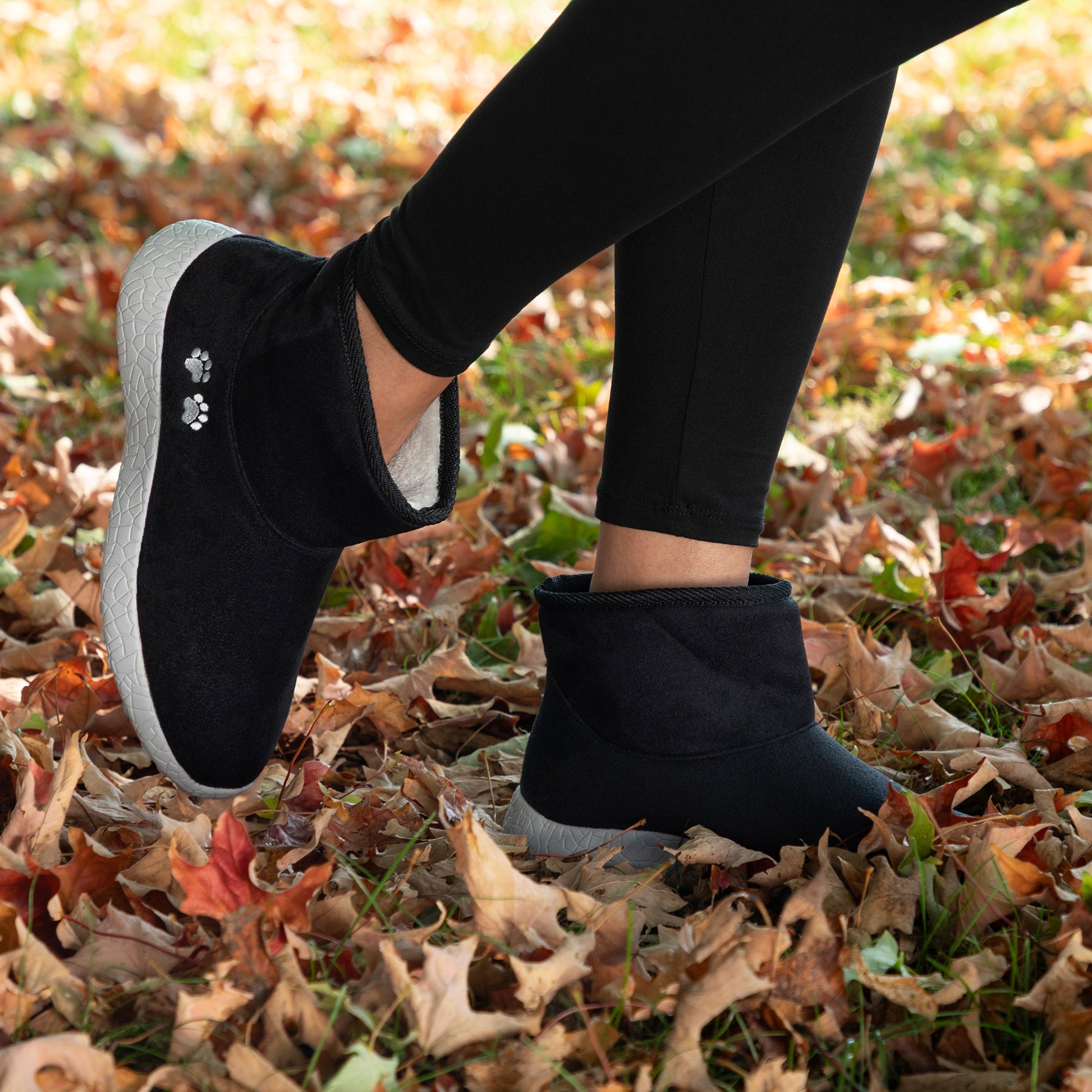 Person wearing black slip-on shoes with gray soles on a leaf-covered ground.
