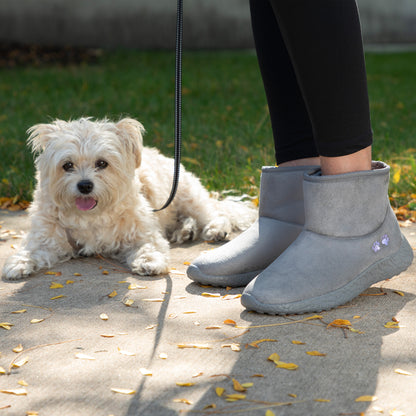 Person wearing gray ankle boots with a small white dog on a leash in a park setting.