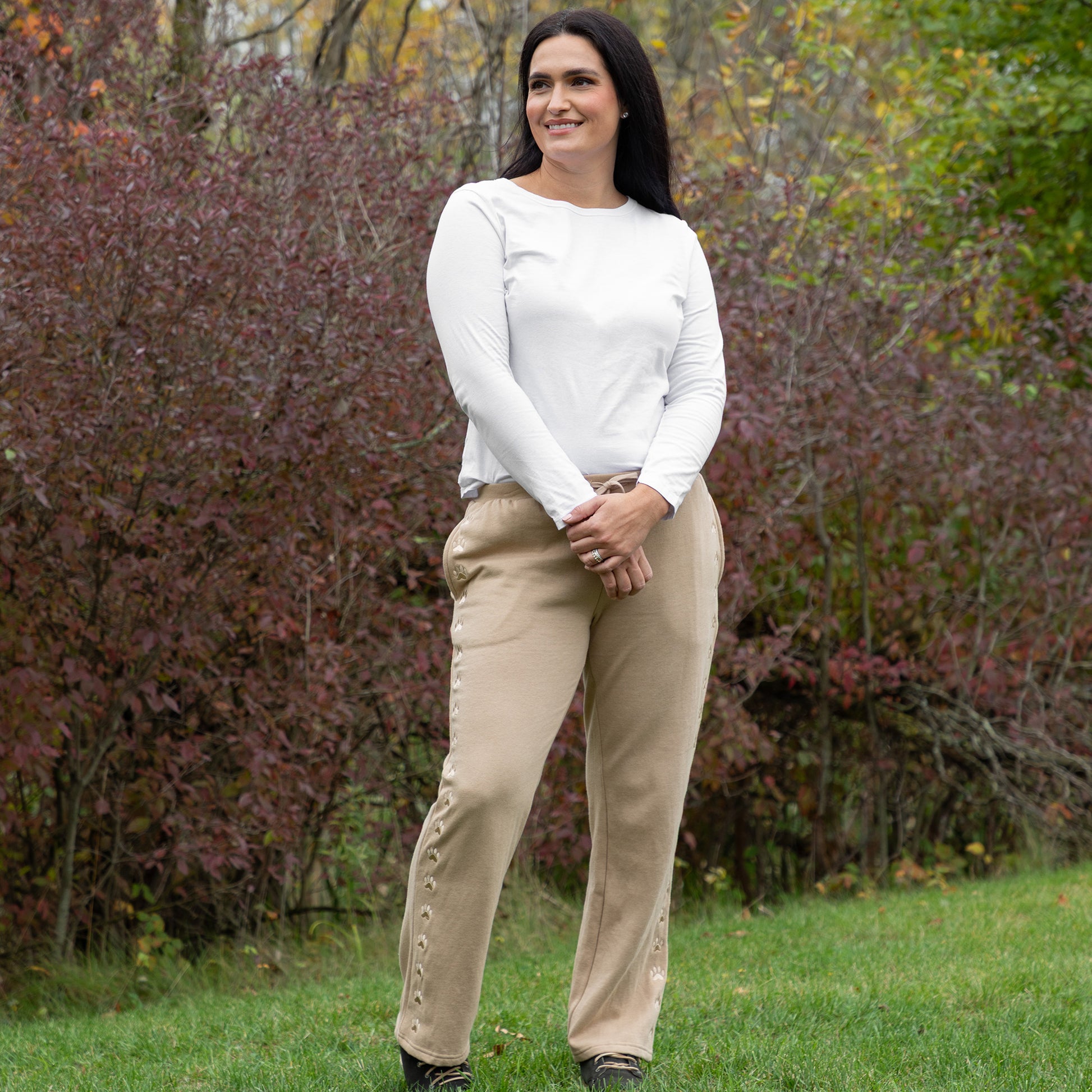 Woman wearing a white long-sleeve top and beige pants standing in a grassy area with trees in the background.