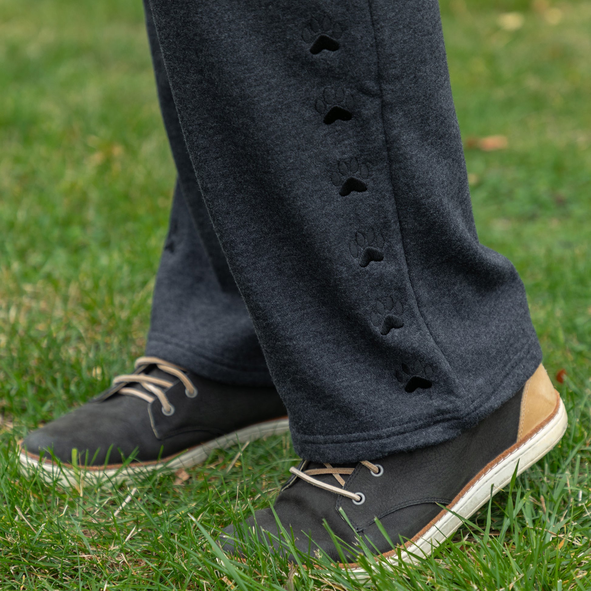 Person wearing dark gray pants with embroidered paw details and black shoes on grass
