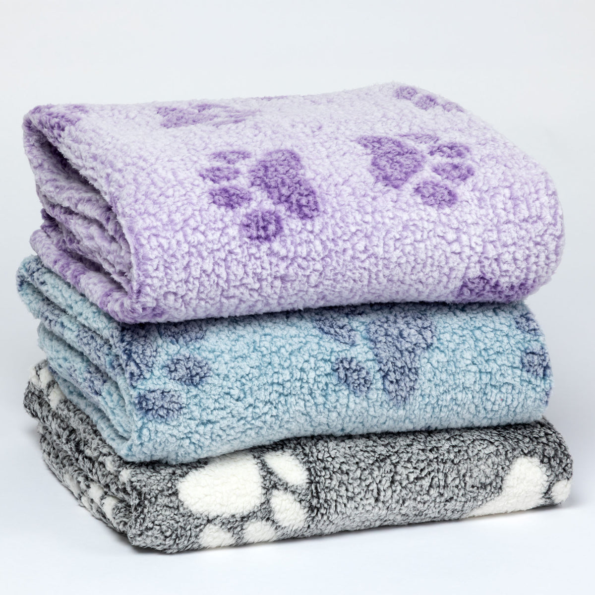 Three folded plush fleece blankets stacked on a white background, each with a purple paw print pattern. The top blanket is lavender, the middle is light blue, and the bottom is gray and white.