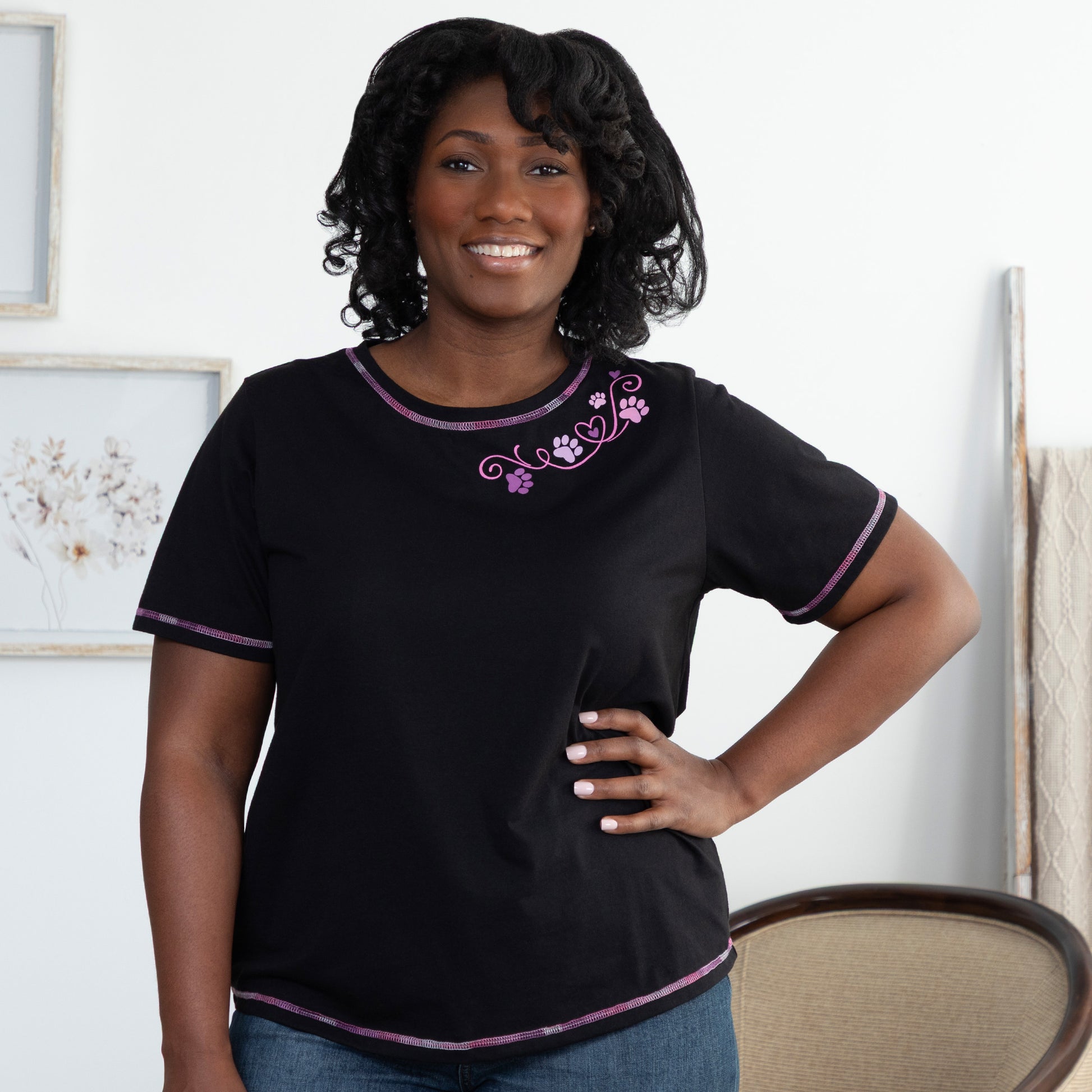 Woman wearing a black t-shirt with purple contrast stitching throughout and a swirling heart and paw print design at the collar, standing in a living room.