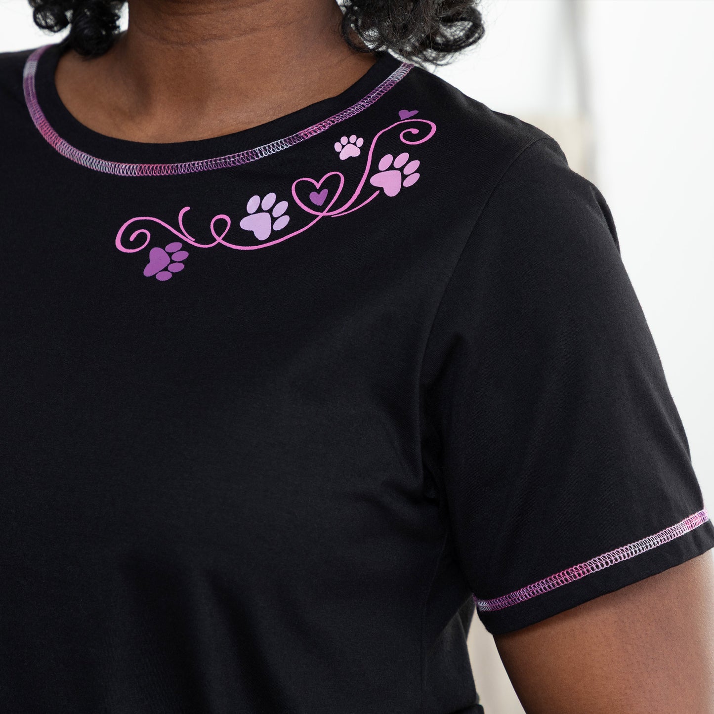 Close up of a black tee with purple contrast stitching throughout and a pink and purple paw and heart design at the neckline.