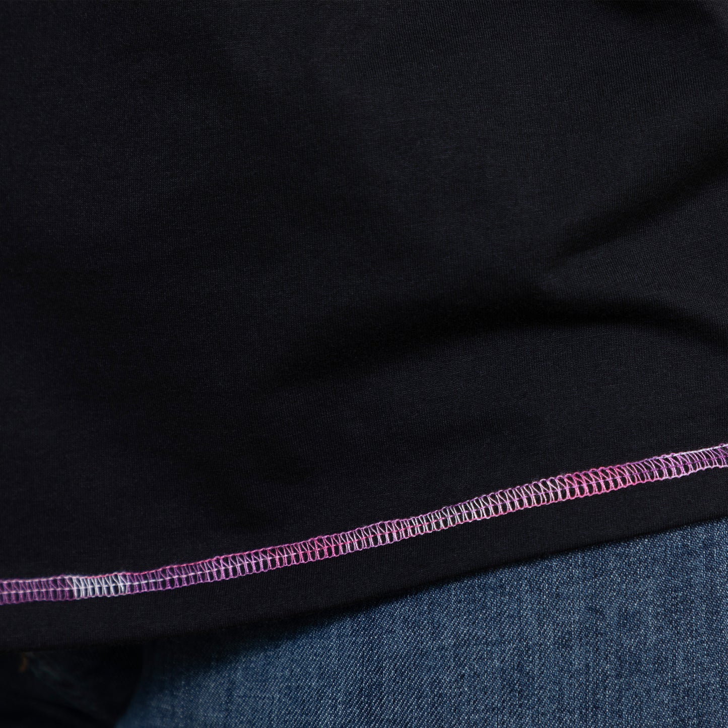 Close-up of black tee fabric with pink and purple contrast stitching detail at hem.