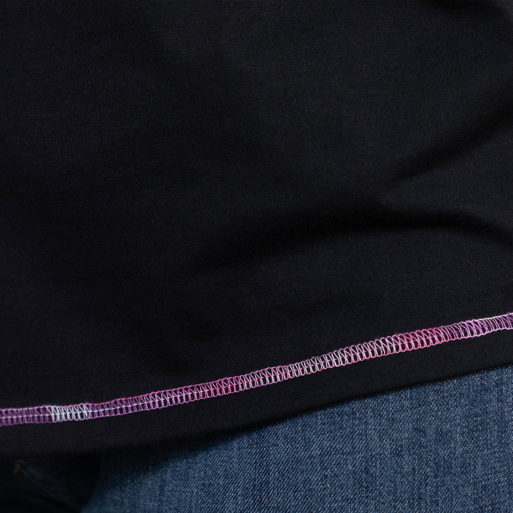Close-up of black tee fabric with pink and purple contrast stitching detail at hem.