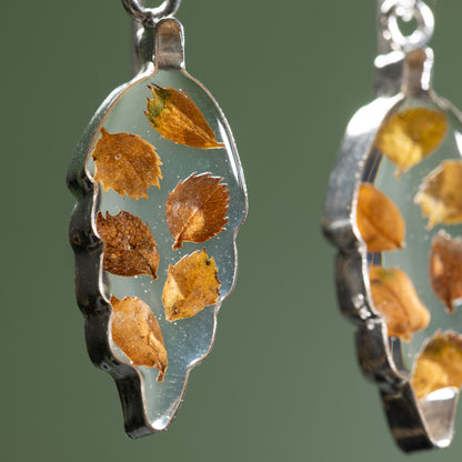 Close up of leaf-shaped earrings with tiny leaves in resin within on a green background