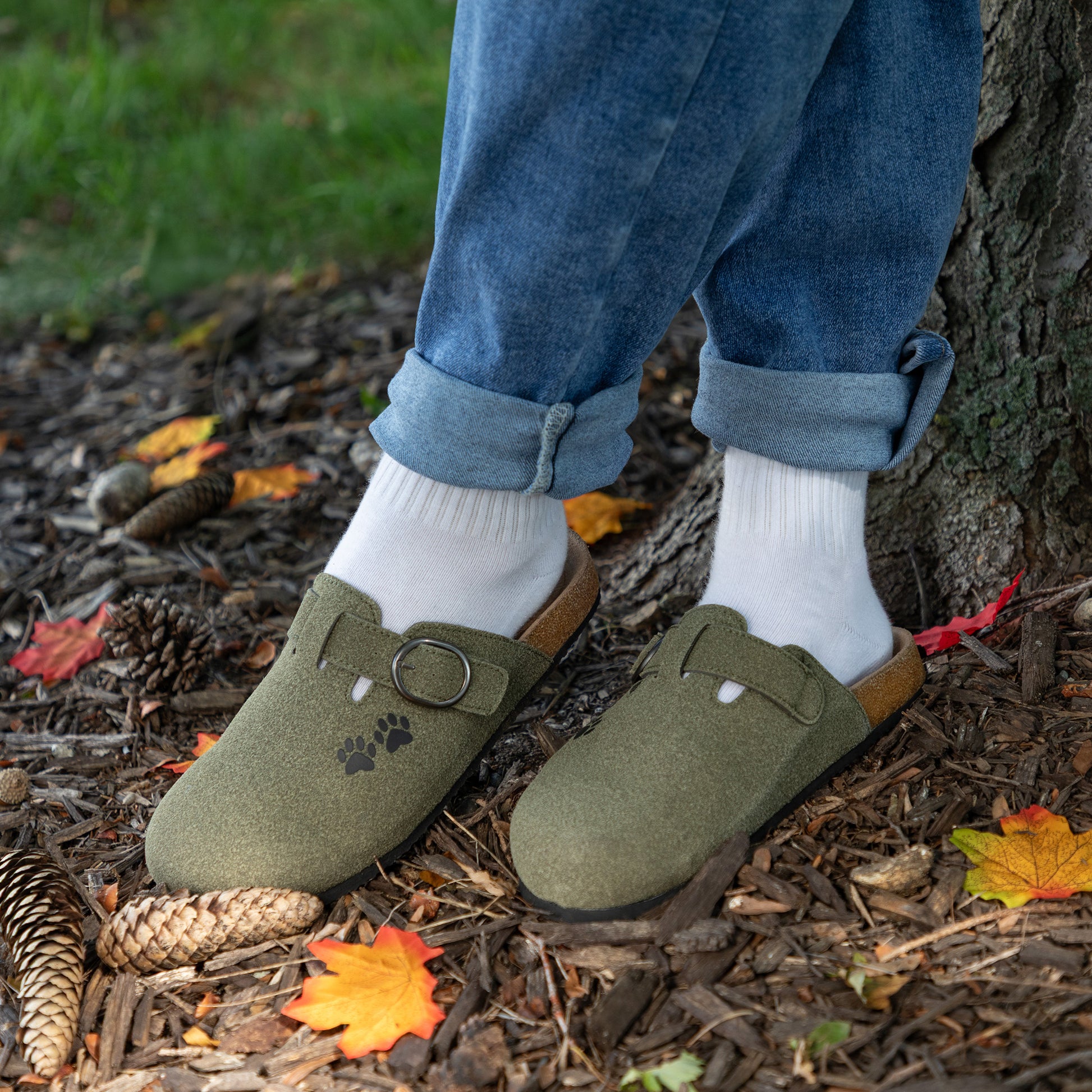 A person wearing olive green clogs with white socks, standing outdoors on a forest floor next to a tree trunk.