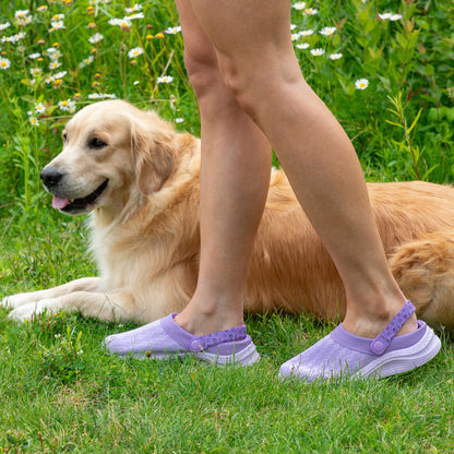 A person wearing light purple slip-on clogs stands on green grass next to a golden retriever dog lying in a field of white flowers.