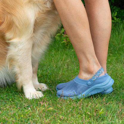 A person wearing light blue tab-style slip-on shoes standing on grass next to a golden retriever dog.
