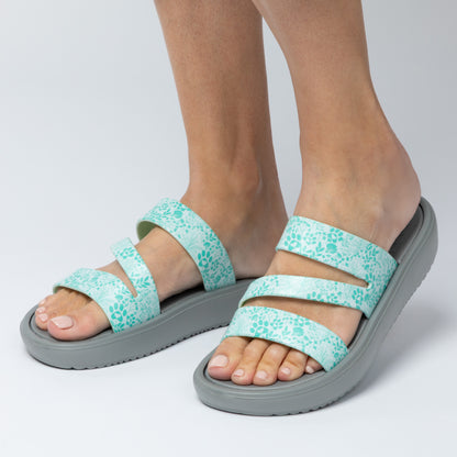 Teal floral sandals with gray soles worn by a person on a light gray background
