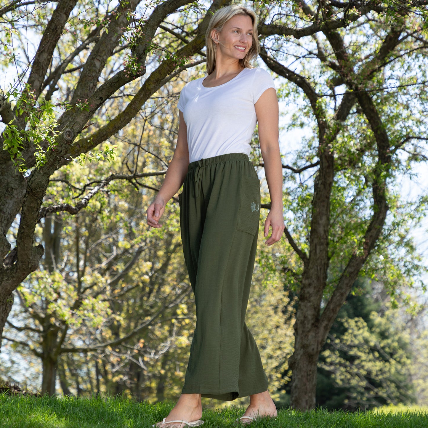 Woman wearing a white top and green pants standing in a park with trees and grass.