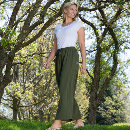 Woman wearing a white top and green pants standing in a park with trees and grass.