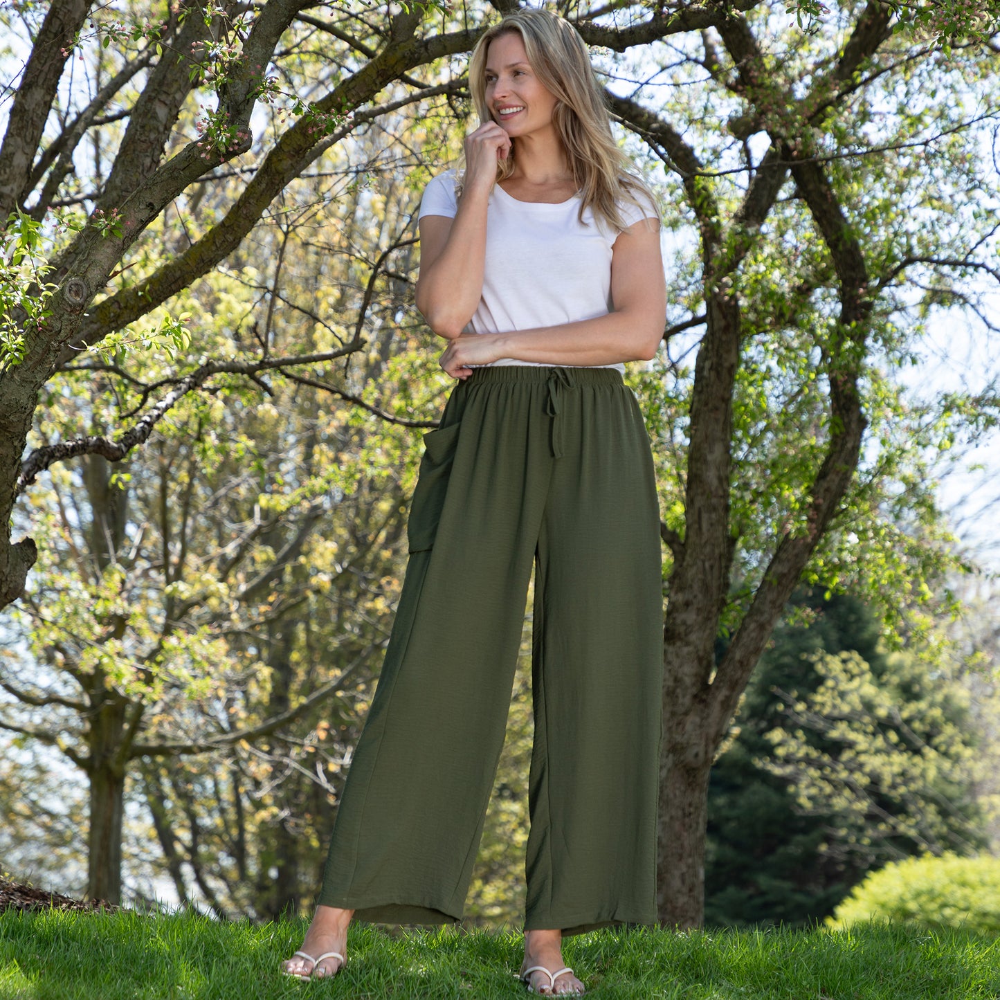 Woman wearing a white top and green wide-leg pants standing in a park.