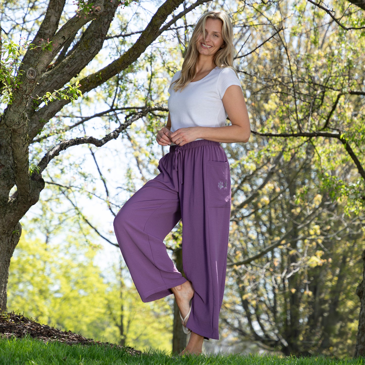 Woman wearing purple pants and a white top standing under a tree in a park.