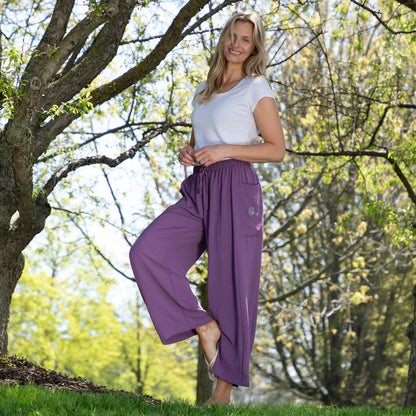 Woman wearing purple pants and a white top standing under a tree in a park.