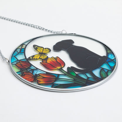 An oval stained-glass-style sun catcher featuring a black dog silhouette, yellow butterflies, red-orange tulips, and blue-green leaves, suspended from a silver chain on a white background.