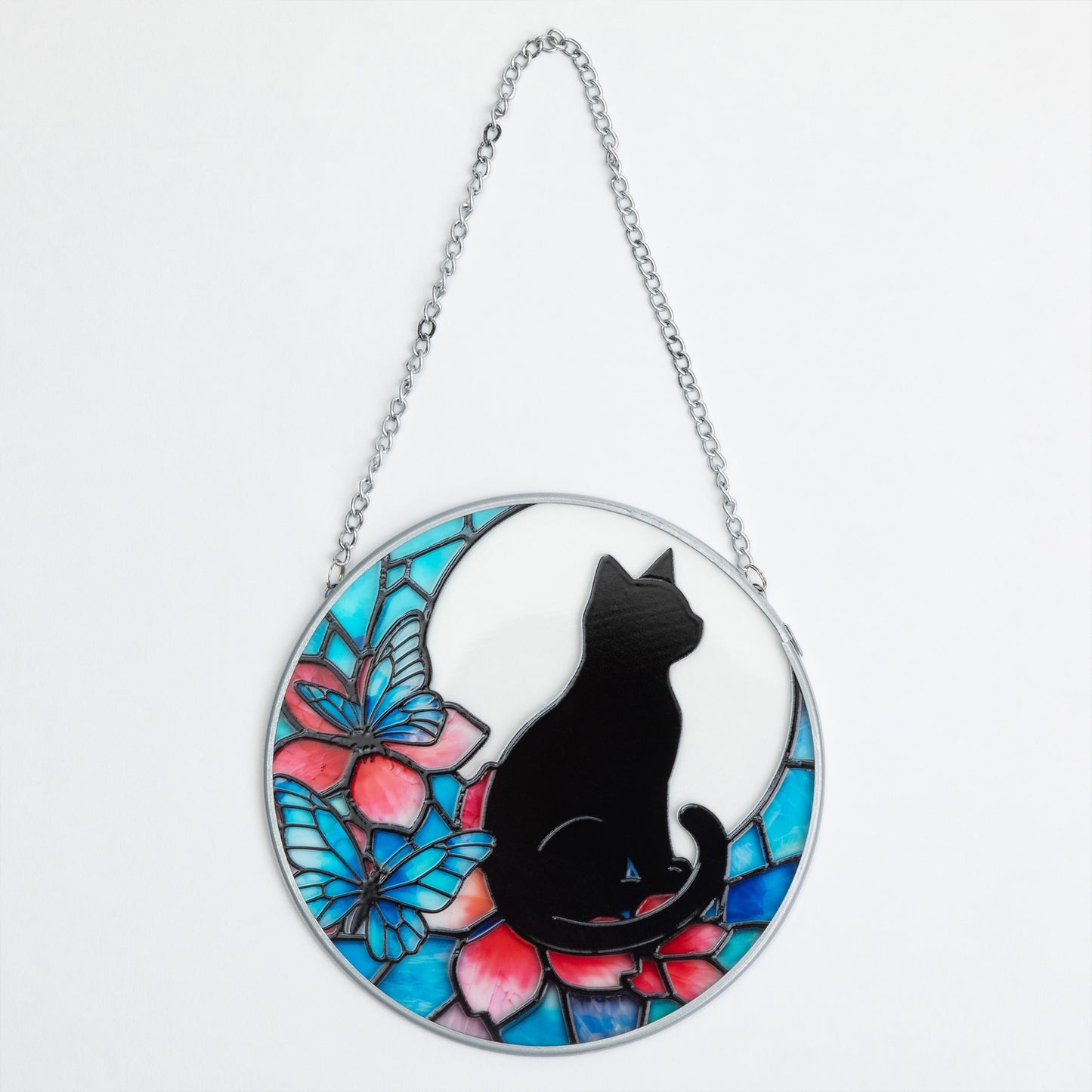 A circular stained-glass-style hanging ornament featuring a black cat silhouette seated in front of a white crescent moon and colorful blue, pink, and teal butterfly and flower motifs, suspended by a silver chain on a white background.