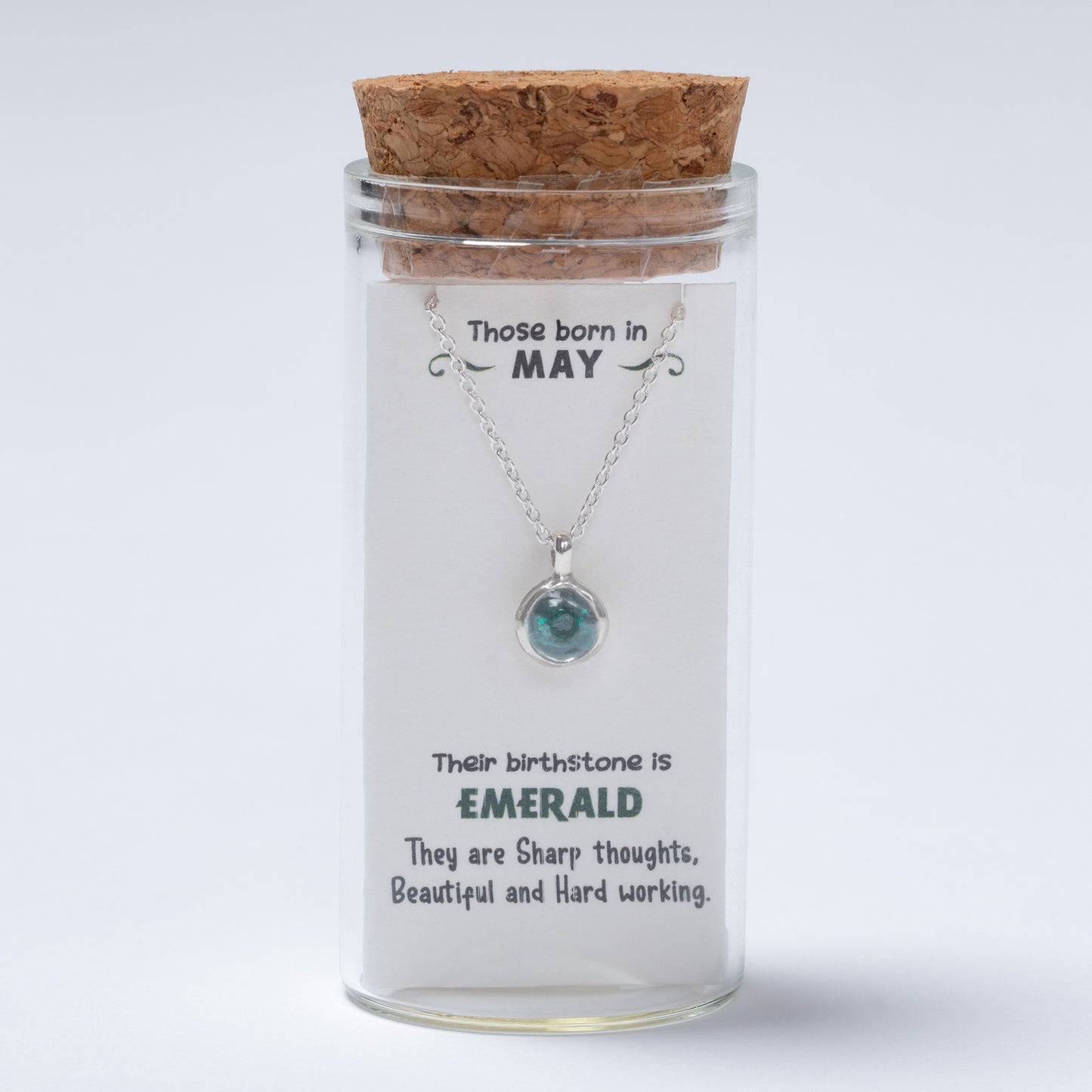 Necklace with a pendant in a glass jar labeled for May birthstone, on a light gray background