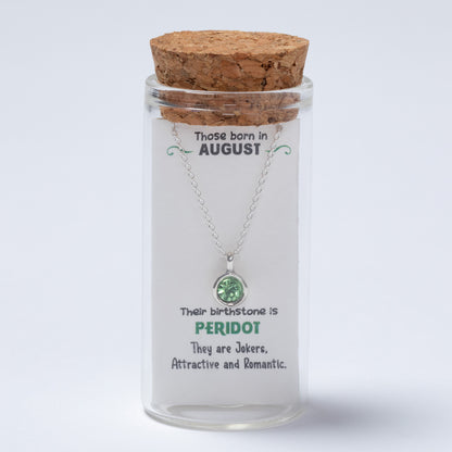 Glass jar with cork lid containing a necklace with a green gemstone, on a light gray background.