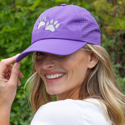 Woman wearing purple cap with embroidered paws on the front, with greenery in the background.