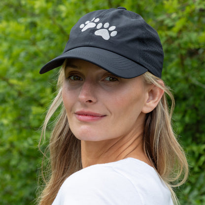 Woman wearing a black hat with two embroidered paw prints on the front, outside in front of greenery.