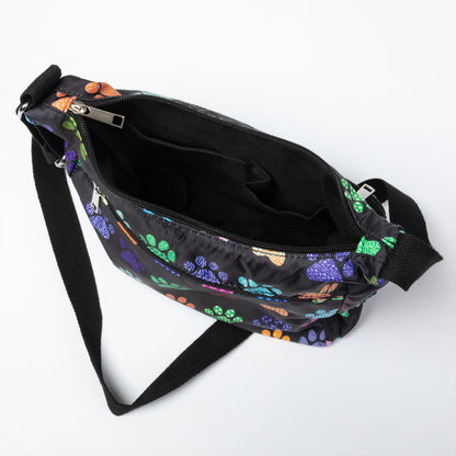 A black nylon crossbody bag with a colorful paw print pattern, shown open to reveal its interior compartments and black strap on a white background.